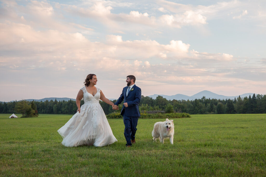 Wedding couple runs carefree through a field at sunset with their dog