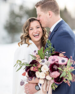 A groom kisses his bride on the forehead while she laughs and looks at the camera. She's holding a pink and burgundy bridal bouquet.