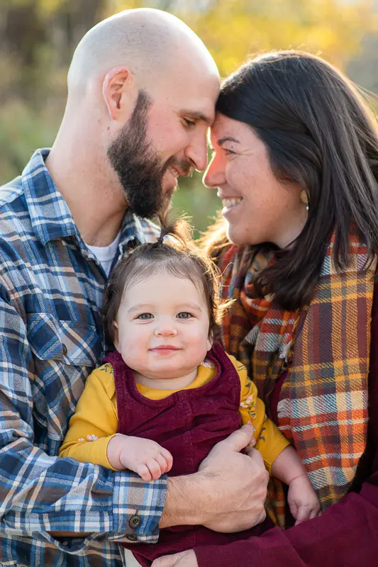 Mom and Dad hold baby with heads together, baby girl looks at camera