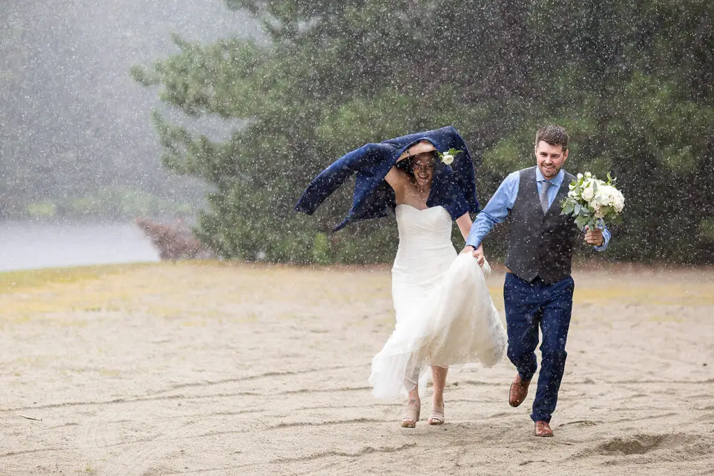 A bride and groom run towards their vermont wedding photographer on a beach trying not to get too wet in the rain. The bride is covering her head with the groom coat. The groom. Is holding up the brides dress so not to get sandy