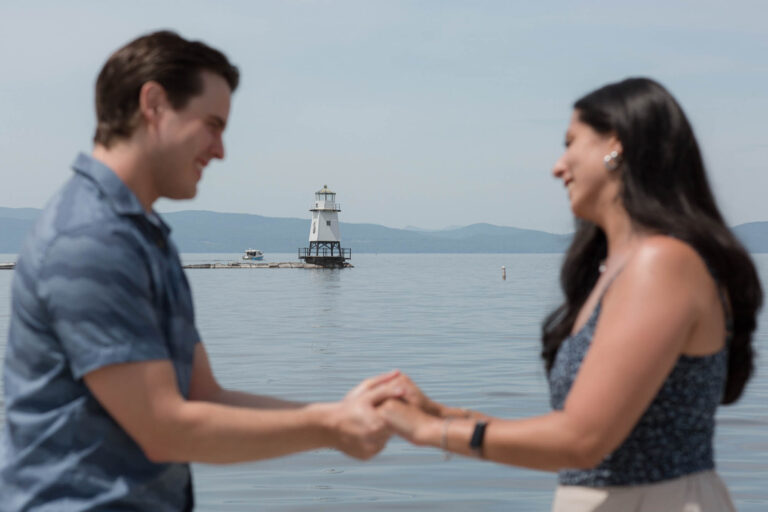 The lighthouse is in focus in the background as a couple gets engaged on the fishing pier in Burlington, Vermont. It's a beautiful summer morning with a blue shy and lake Champlain is calm.