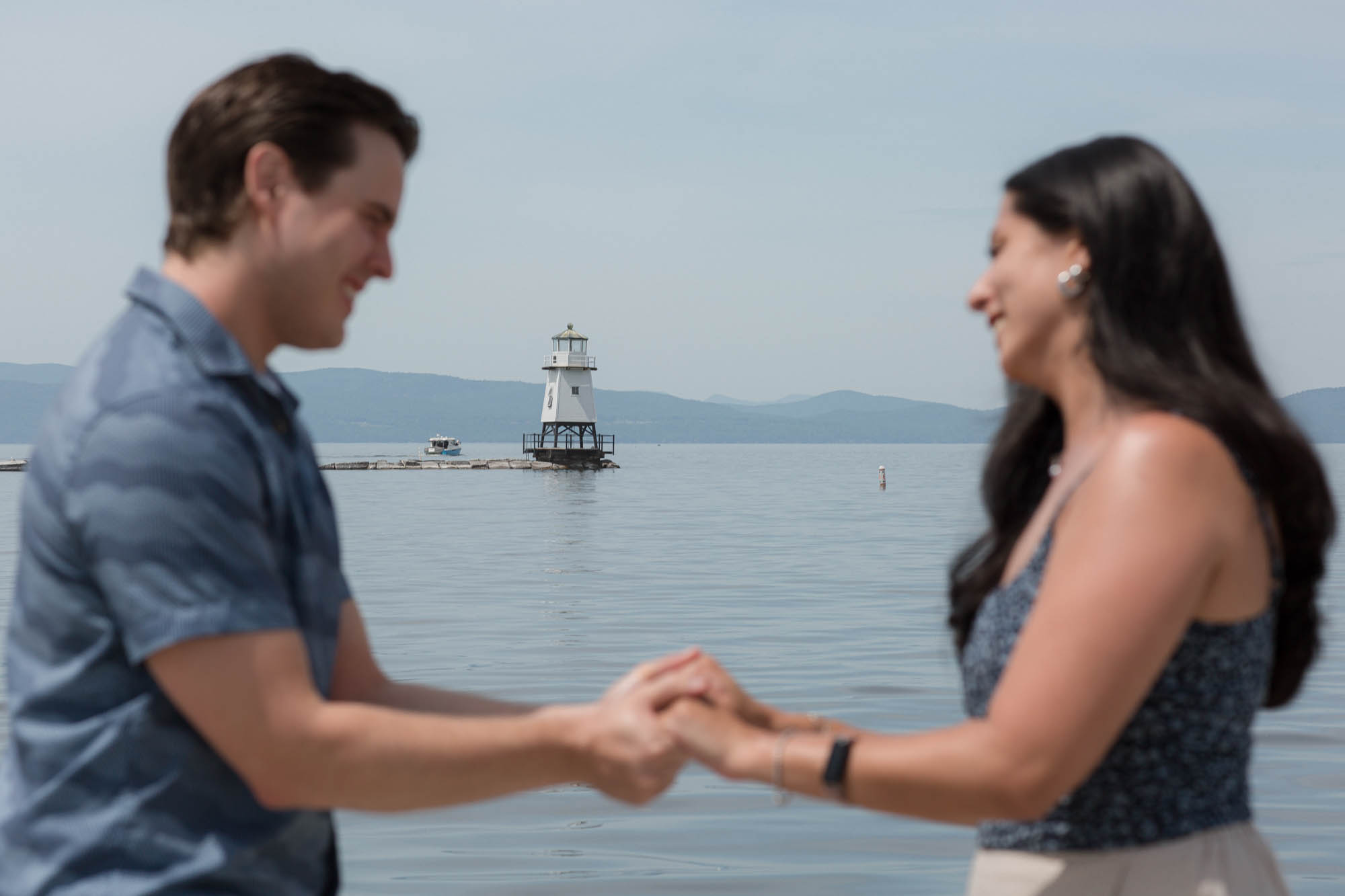 The lighthouse is in focus in the background as a couple gets engaged on the fishing pier in Burlington, Vermont. It's a beautiful summer morning with a blue shy and lake Champlain is calm.